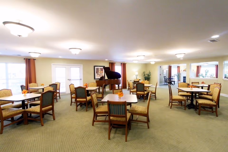 A spacious dining room with multiple square tables and wooden chairs arranged neatly. Each table has a small floral centerpiece. The room has beige walls, green carpet, and several ceiling lights. There is a grand piano against the far wall, and large windows with orange curtains letting in natural light.