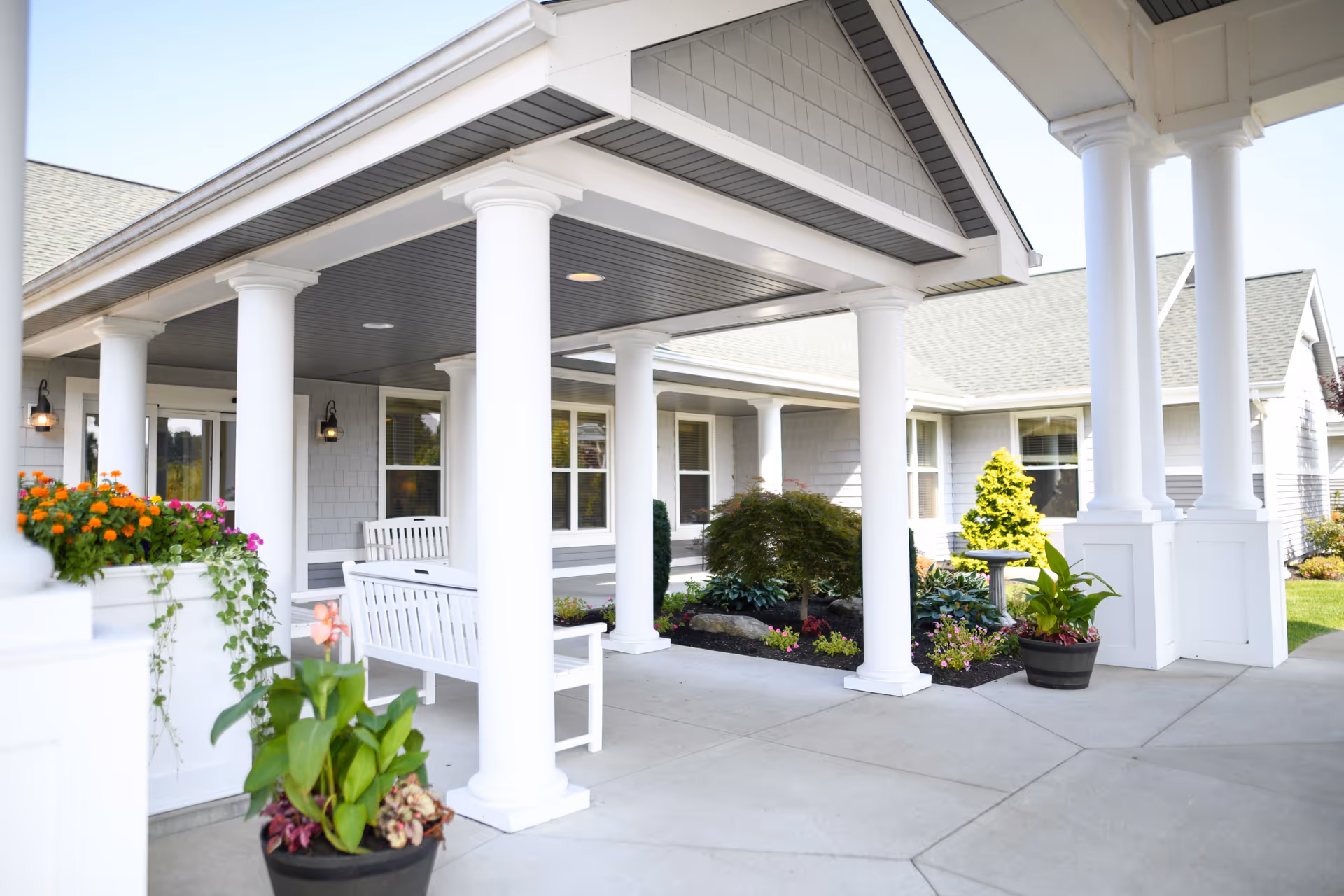 Covered outdoor patio area with white columns and benches, surrounded by potted plants and flower beds, attached to a light gray building with multiple windows.