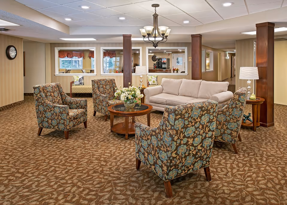 A cozy living room area with a beige sofa, four floral-patterned armchairs arranged around a round wooden coffee table with a flower arrangement. The room has carpeted flooring with a leaf pattern, beige walls, wooden columns, a chandelier, and a table lamp on a side table. Windows in the background show another room with seating and a clock on the wall.