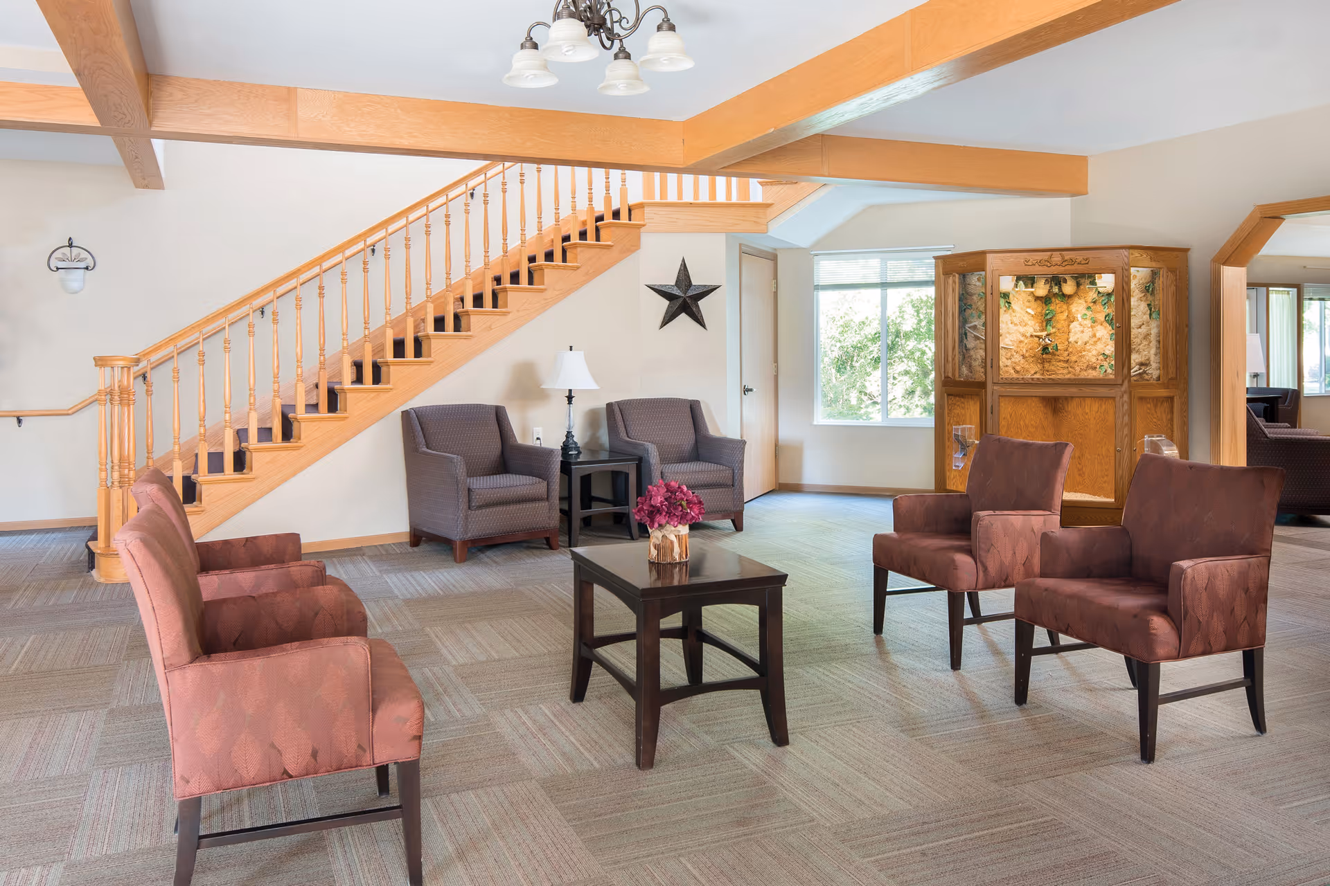A bright and spacious living room area with a wooden staircase along the left wall. The room features a mix of upholstered chairs in muted red and gray colors arranged around a dark wooden coffee table with a vase of flowers. There is a wooden display cabinet with decorative items near a large window letting in natural light. The ceiling has wooden beams and a chandelier with multiple lights.
