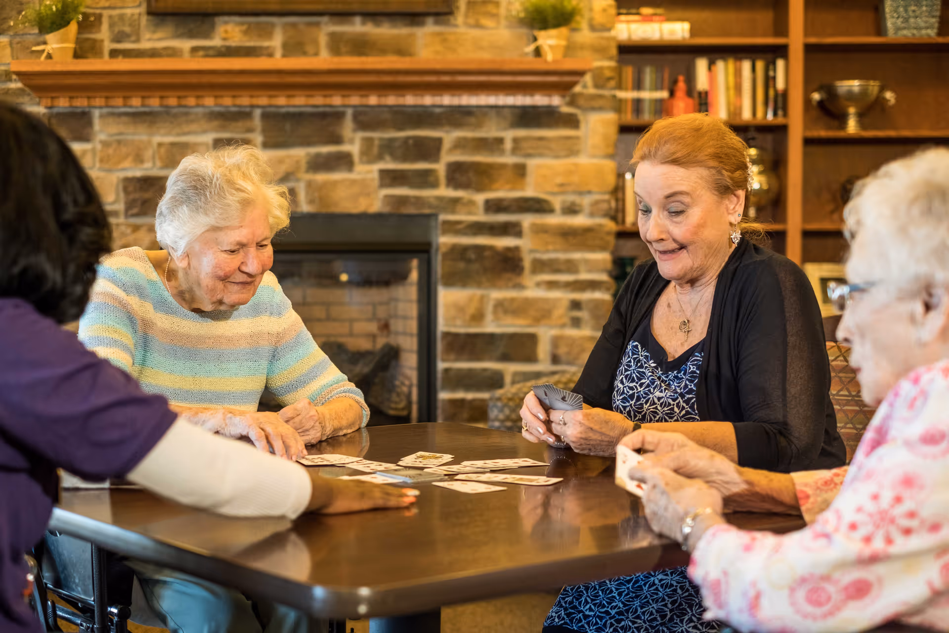 Four elderly women sitting around a table playing cards in a cozy room with a stone fireplace and wooden shelves filled with books and decorative items.