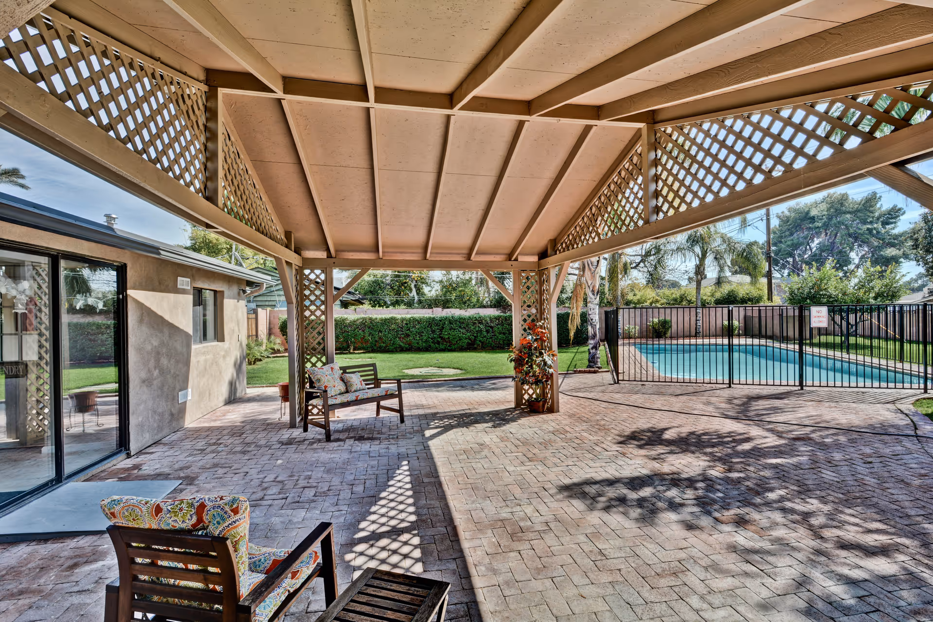Covered patio with seating overlooking a fenced swimming pool and landscaped yard.