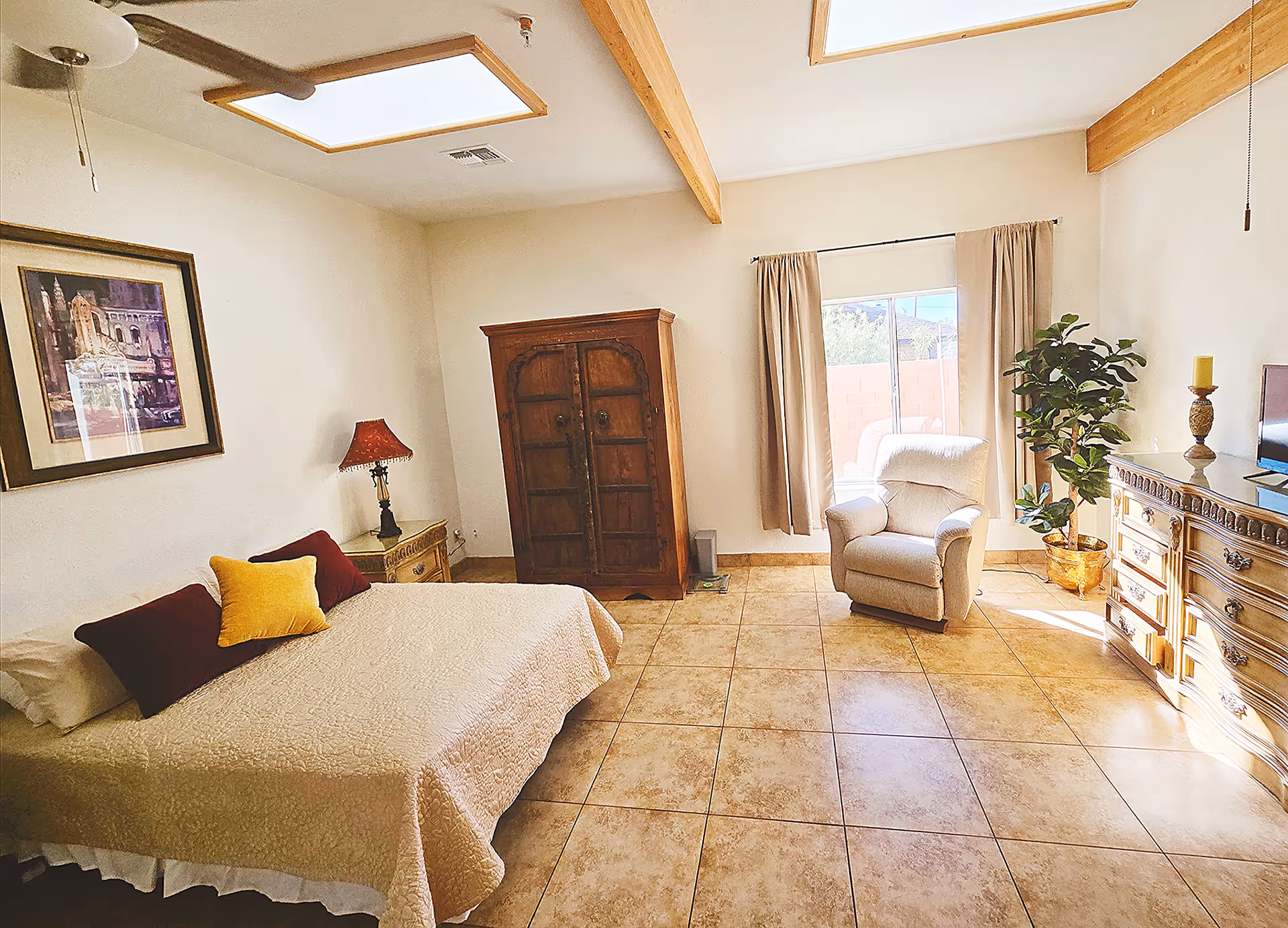 A bright bedroom with beige tiled floor and white walls featuring wooden ceiling beams. The room contains a bed with beige quilt and three pillows in red and yellow, a wooden nightstand with a red lamp, a large wooden armoire, a beige armchair near a window with beige curtains, a potted plant, and a wooden dresser with a TV and decorative items on top.