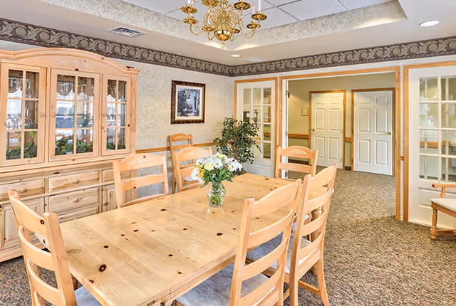 A well-lit dining room with a large wooden table surrounded by six matching wooden chairs. A vase with white flowers is placed in the center of the table. Behind the table is a wooden china cabinet with glass doors displaying glassware. The room has patterned wallpaper with a decorative border near the ceiling, a chandelier, and a framed picture on the wall. There are double doors leading to another room with beige walls and carpeted floors.