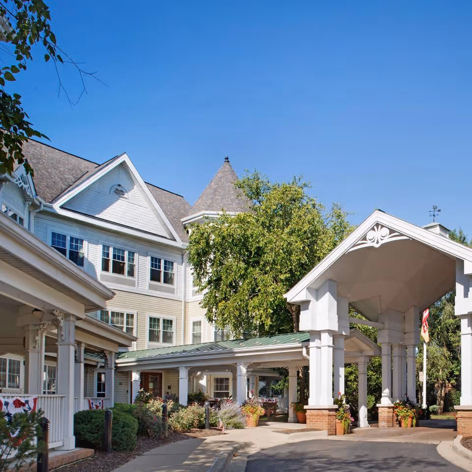 Exterior view of a senior living facility with a covered entrance supported by white columns. The building has multiple windows, a peaked roof, and decorative architectural details. There are trees and plants around the entrance, and the sky is clear and blue.