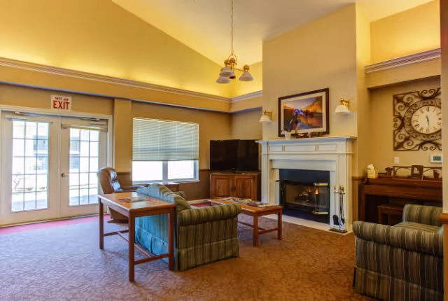 A cozy living room with a striped sofa and armchair, a wooden coffee table, a fireplace with a framed picture above it, a flat-screen TV on a wooden cabinet, a large clock on the wall, and a door with a 'Not an Exit' sign leading outside.