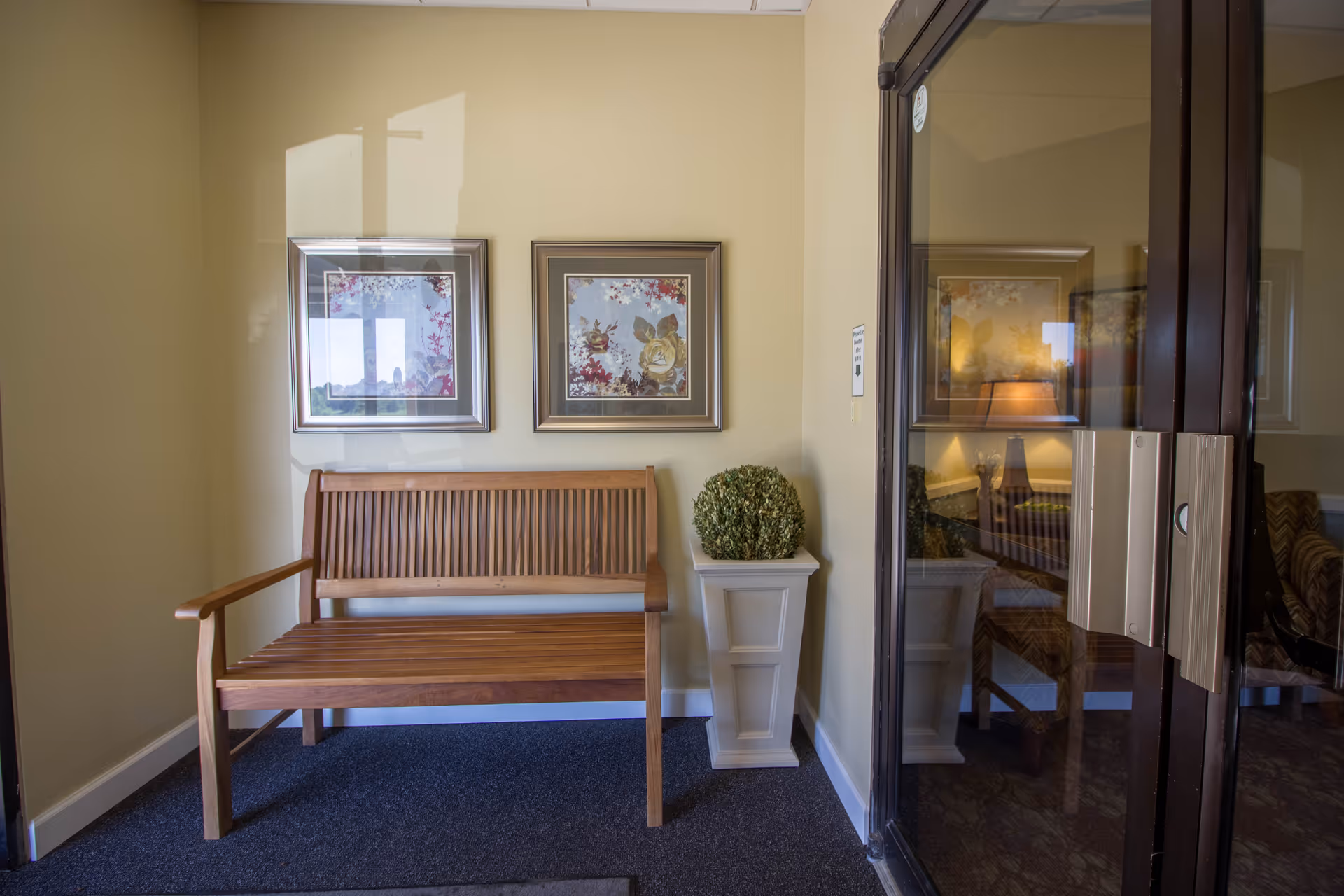 Wooden bench and potted topiary beneath two framed floral prints next to a glass entry door reflecting a seating area.