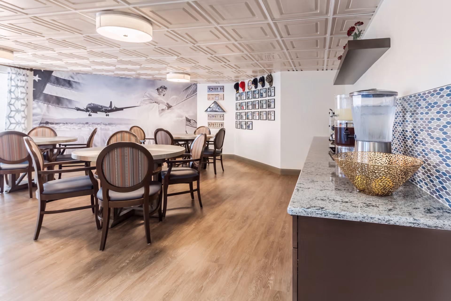 Dining area with round tables and chairs, a beverage station on a countertop, and a large aviation-themed mural on the wall.