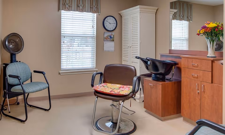 A senior living facility hair salon room with a salon chair featuring a floral cushion, a hair washing station with a black sink, a wooden cabinet with drawers, a vase of colorful flowers on the counter, a blue cushioned chair, a hair dryer, a window with blinds and a valance, a wall clock, and a calendar.