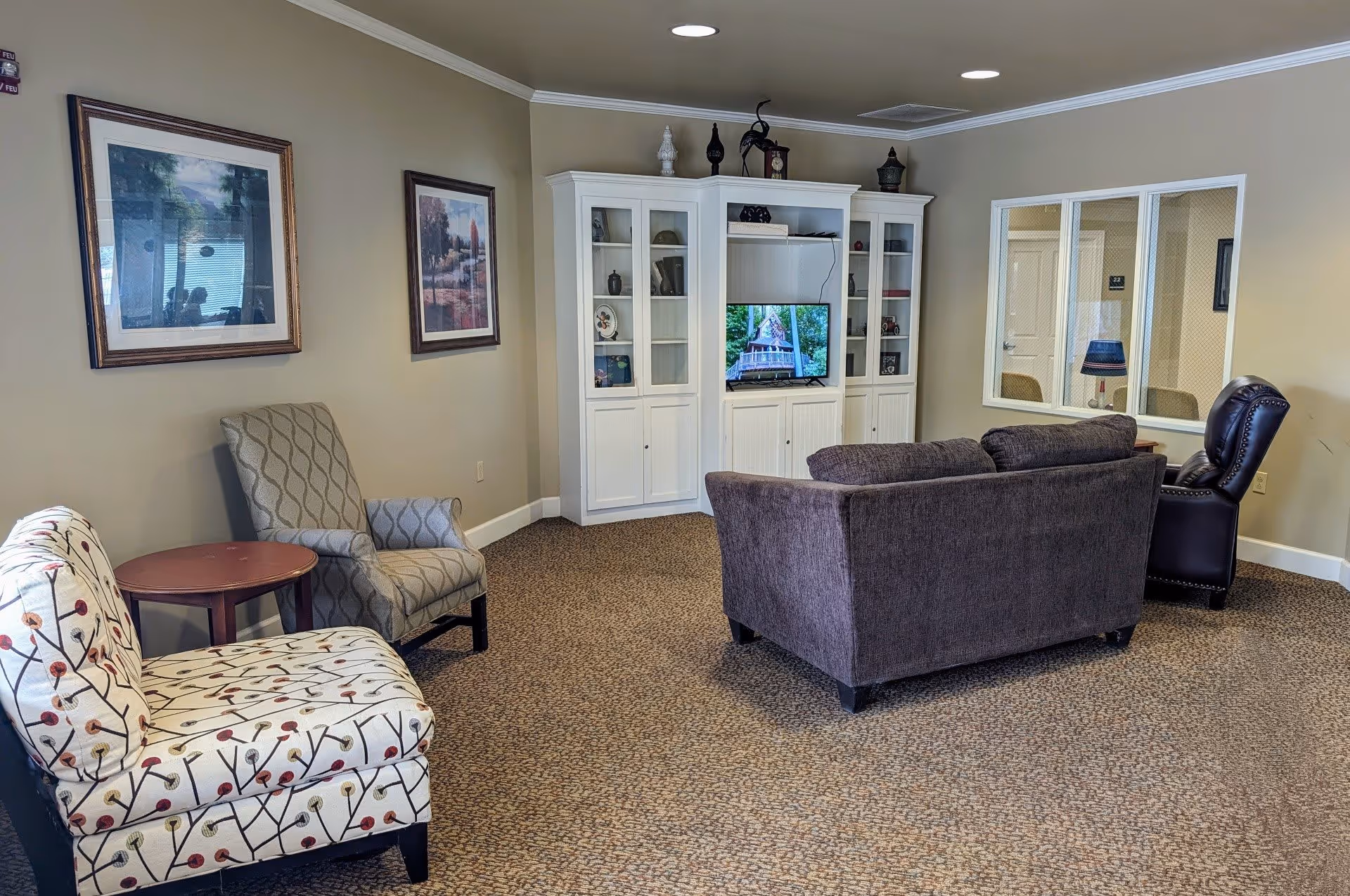 A cozy living room area with a patterned armchair, a gray armchair, a dark gray sofa, and a black leather recliner. There is a white entertainment center with glass cabinets and a TV displaying an image of a house. The walls are beige with two framed pictures, and there is a window showing an adjacent room with a lamp and chairs.