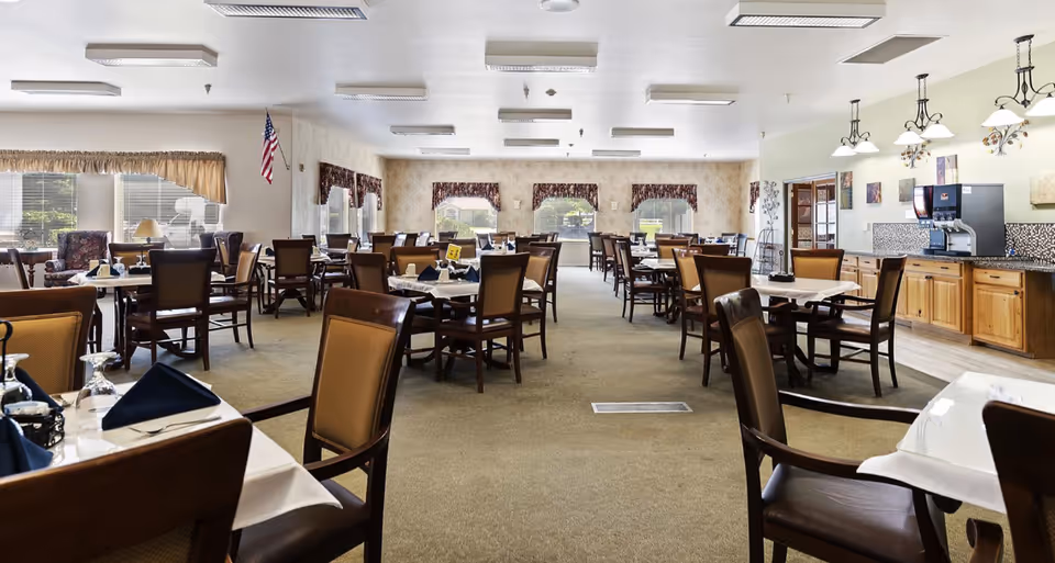 A spacious dining room in Autumn Wind Assisted Living with multiple tables and chairs arranged neatly. The tables are set with white tablecloths, napkins, and glassware. Large windows with valances allow natural light to fill the room. There is a beverage station with a soda dispenser on the right side of the room, and the walls are decorated with light-colored wallpaper and framed artwork.