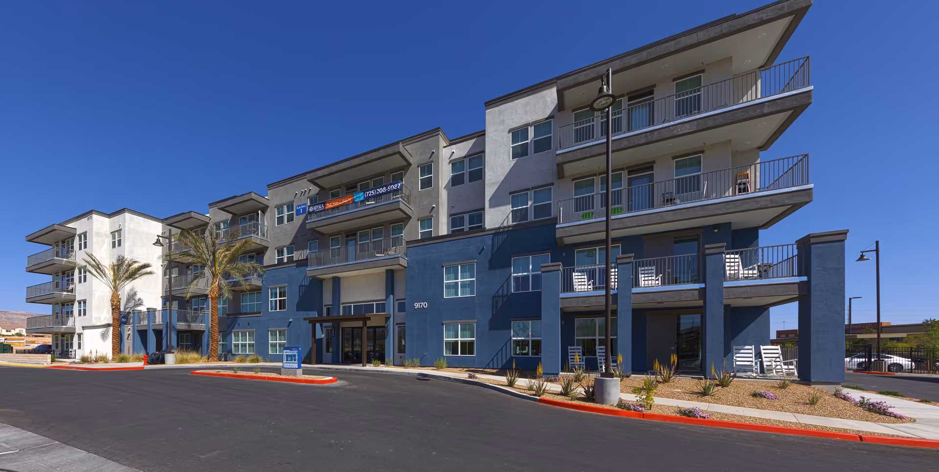 Exterior view of a modern four-story residential building with balconies, palm trees, and a clear blue sky. The building has a mix of white and blue-gray walls and a paved driveway in front.