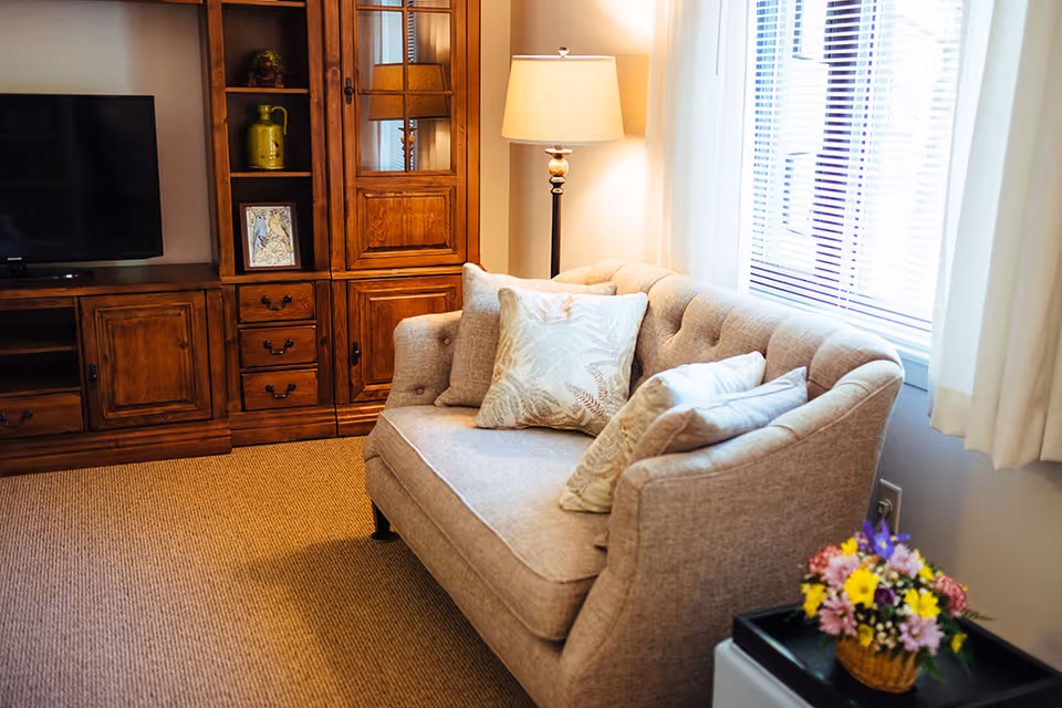 A cozy living room area with a beige tufted sofa adorned with three patterned cushions. Next to the sofa is a floor lamp with a beige shade, and a window with white blinds and curtains. On the right side, there is a small table with a basket of colorful flowers. On the left side, there is a wooden entertainment center with a flat-screen TV, decorative items, and framed artwork.