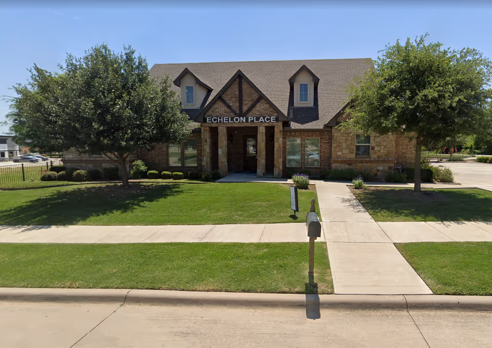 Brick and stone assisted living building front with an 'ECHELON PLACE' sign, lawn, two trees, a sidewalk and a mailbox.