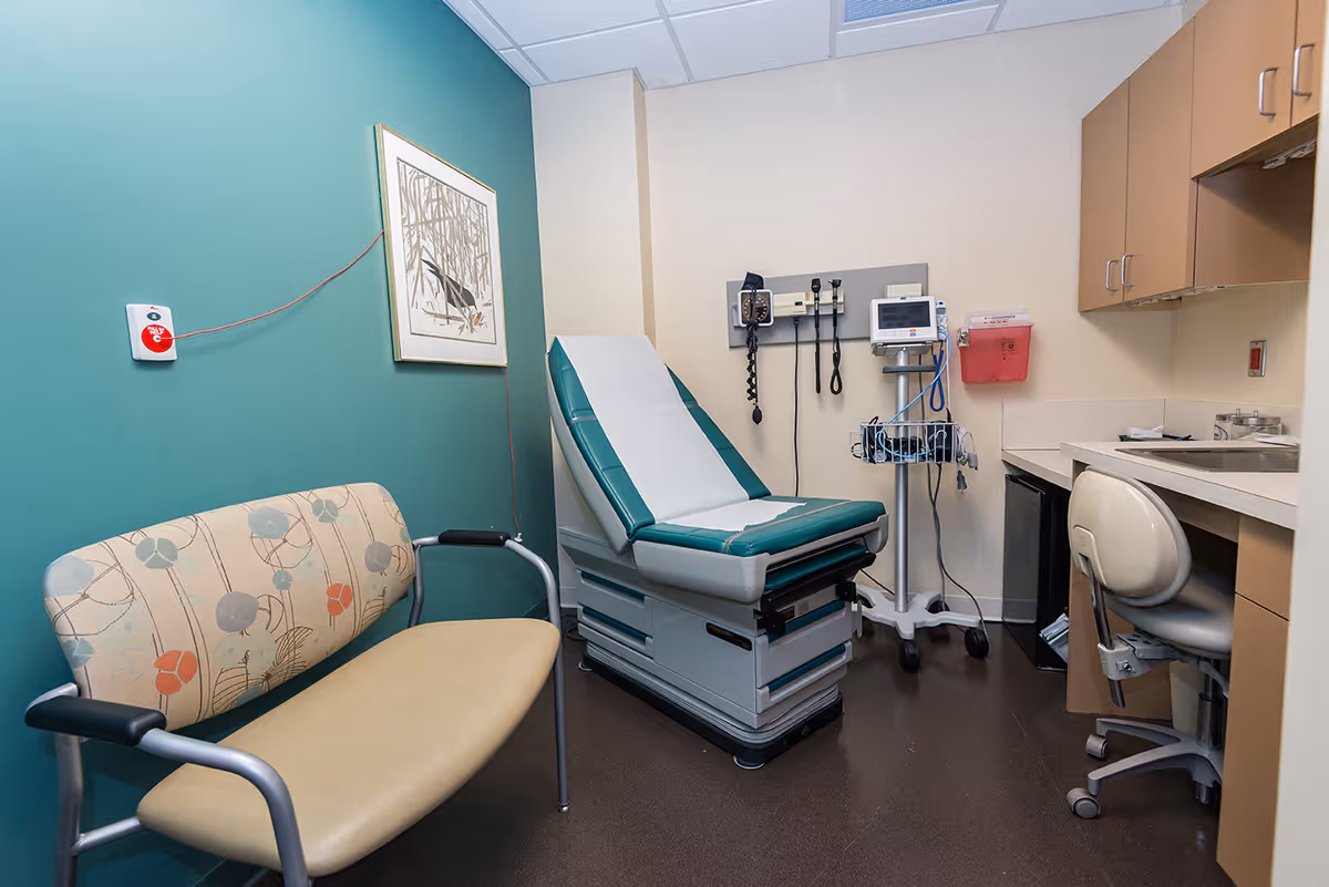 A medical examination room with a teal accent wall, a patterned bench with armrests, an examination table covered with white paper, medical instruments mounted on the wall, a blood pressure monitor on a stand, a small refrigerator, a desk with a chair, and cabinets above the desk.