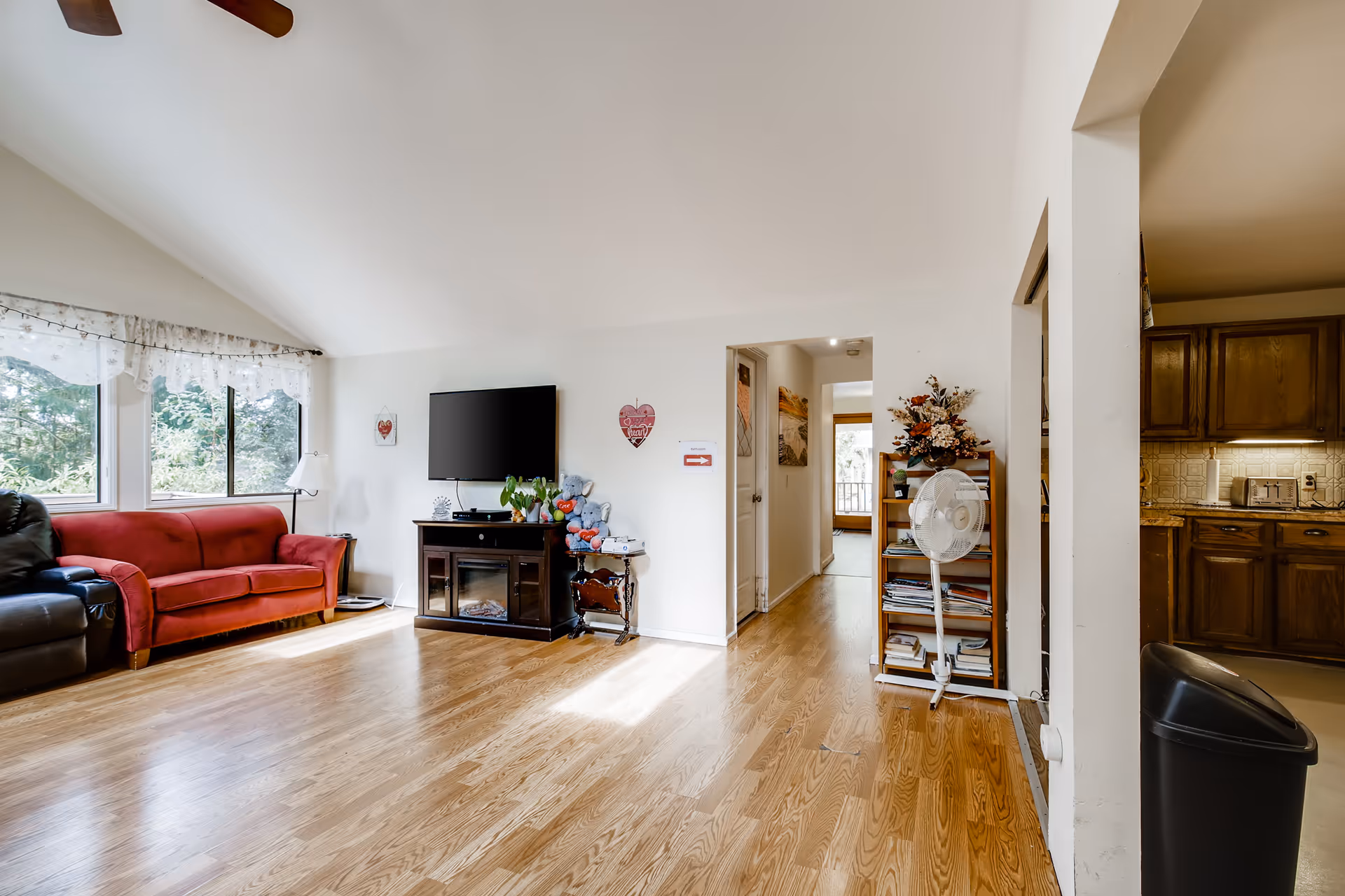 A bright living room with wooden flooring, a red sofa, a black recliner chair, a wall-mounted flat-screen TV above a dark wooden cabinet, and a small side table with teddy bears. There is a window with sheer white curtains letting in natural light. A hallway leads to other rooms, and a wooden bookshelf with a standing fan is visible near the entrance to a kitchen area with wooden cabinets and a trash bin.