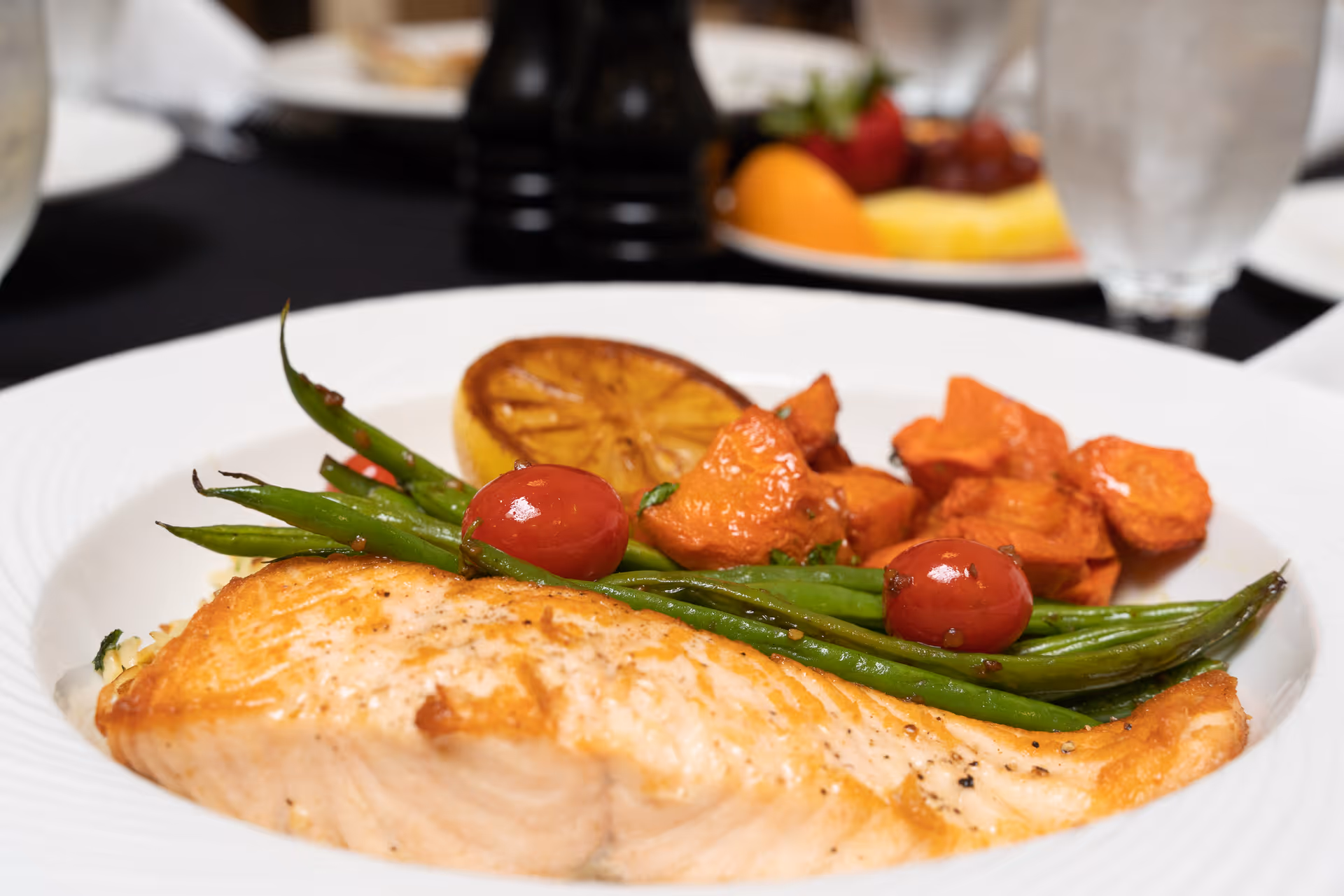 Close-up of a plated salmon entrée with green beans, cherry tomatoes, roasted potatoes and a grilled lemon on a white plate.