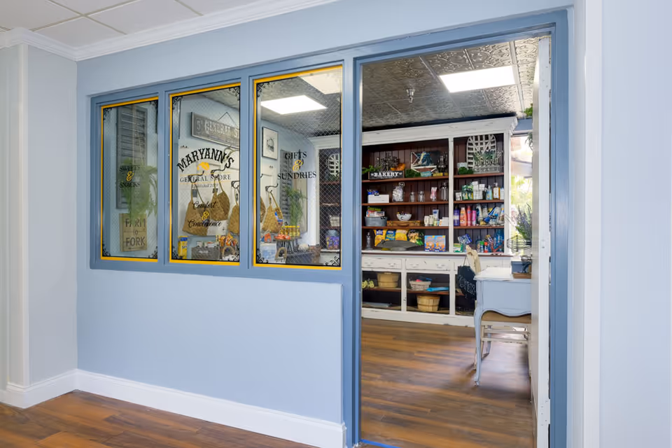 Interior view of a small general store area inside a facility, seen through a blue-framed window and doorway. The store has shelves stocked with various snacks, gifts, and sundries, with a sign that reads 'Maryann's General Store'. The walls are painted light blue and the floor has wood-style flooring.