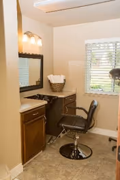 Small salon-style room with a hairstyling chair in front of a mirror, countertop with sink, and a window with blinds.