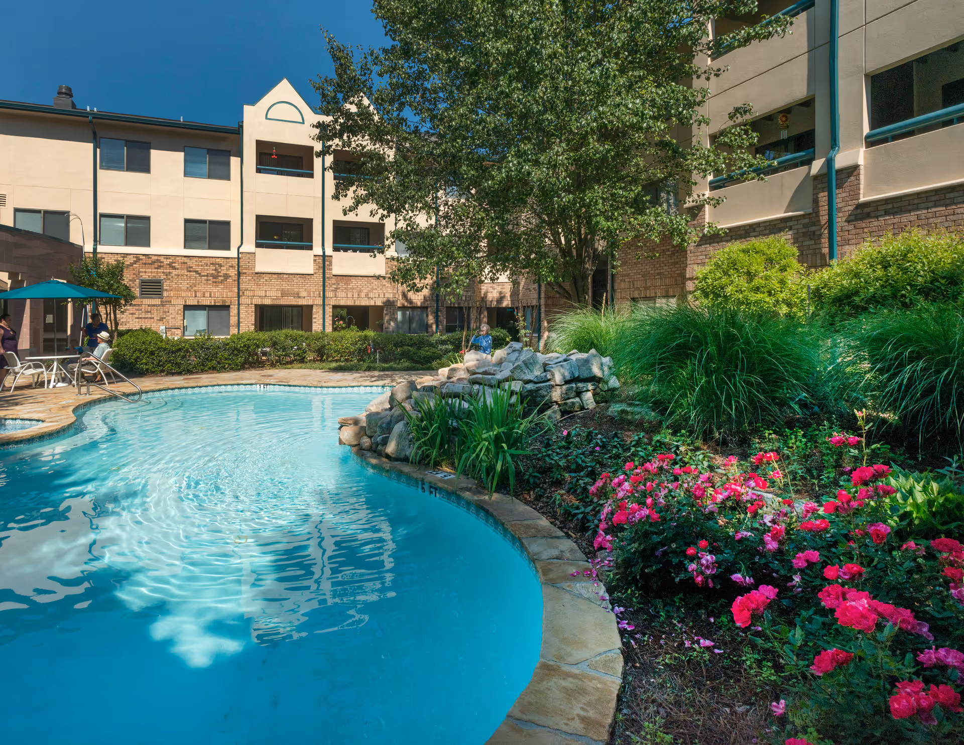 Outdoor courtyard showing a swimming pool, rock waterfall, landscaped flower beds and a multi-story senior living building in the background.