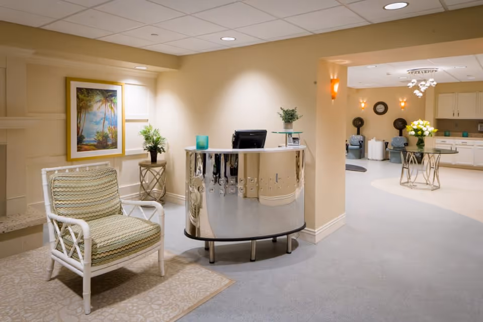 A bright and clean reception area in a senior living facility featuring a modern curved reception desk with a computer, a white wicker chair with patterned cushions, a small side table with a potted plant, and a framed tropical painting on the wall. In the background, there is a spacious room with salon hair dryers, a round glass table with a flower arrangement, and a kitchenette area with cabinets and a countertop.