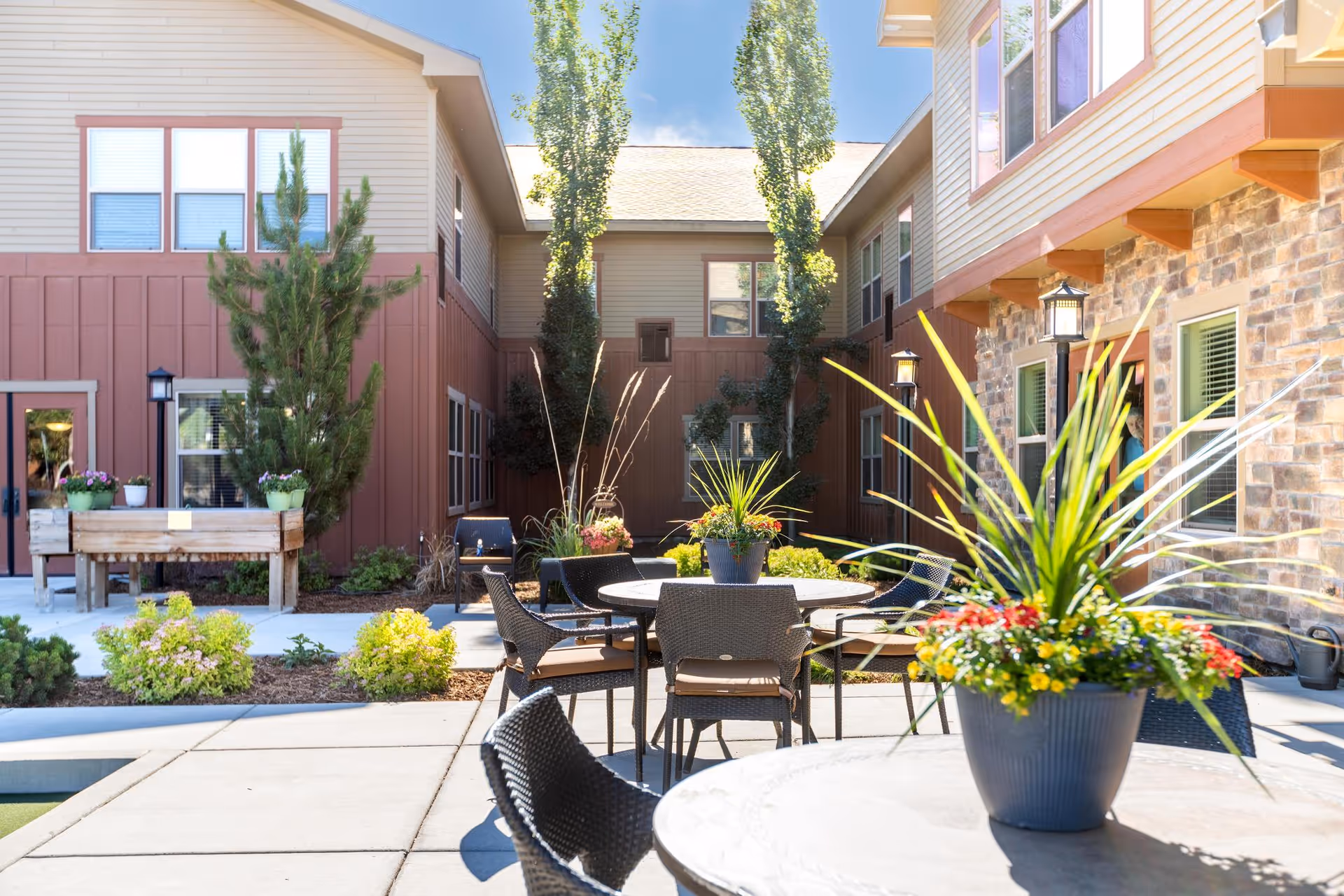 Outdoor courtyard area at Summit Senior Living with round tables and chairs, potted plants with colorful flowers on the tables, surrounded by buildings with windows and a clear blue sky above.