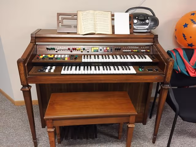 A vintage wooden electric organ with two keyboards, colorful buttons, and switches. On top of the organ is an open sheet music book and a small black stereo. A wooden bench is placed in front of the organ. To the right, part of a black chair and a colorful ball with stars are visible.