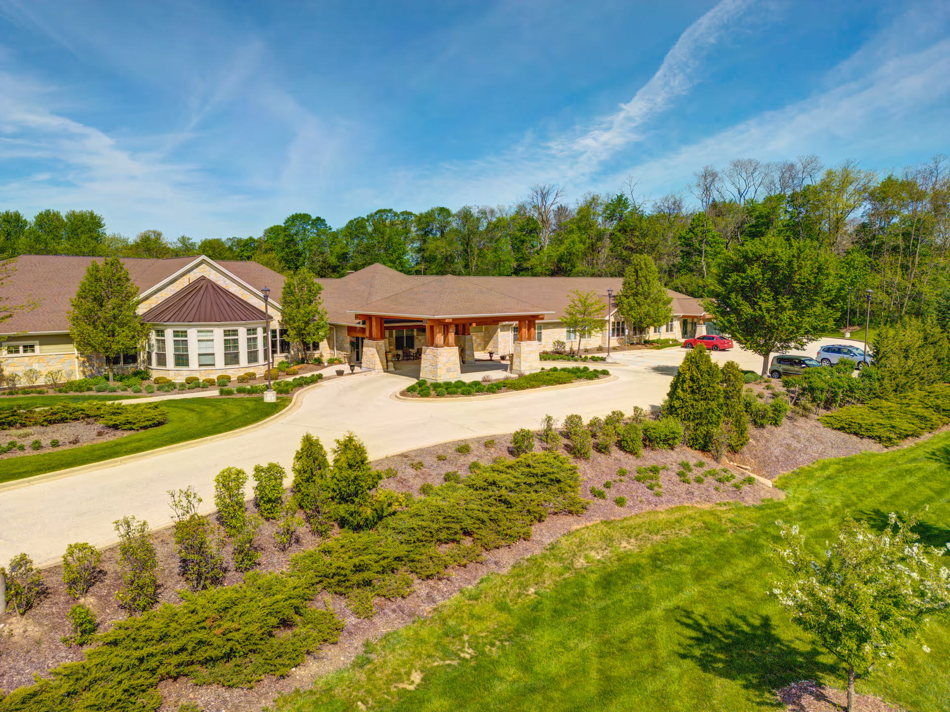 Single-story residential building with a covered porte-cochere entrance, circular driveway, parked cars and manicured landscaping under a blue sky.
