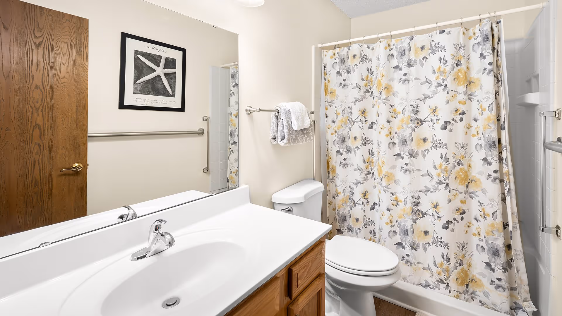 A clean bathroom with a white sink and countertop, wooden cabinets below, a toilet, and a bathtub with a floral shower curtain in shades of yellow and gray. There is a large mirror above the sink, a wooden door, a towel rack with towels, and a framed starfish picture on the wall.