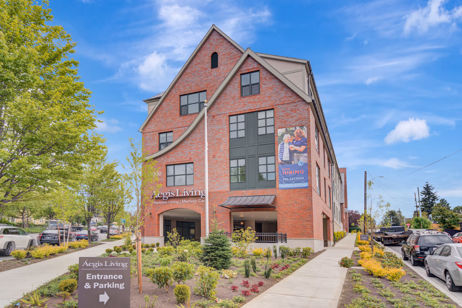 Exterior view of a red brick building with multiple windows under a blue sky with some clouds. The building has a sign that reads 'Aegis Living Assisted Living | Memory Care' and a banner advertising hiring. There is a landscaped area with small trees and bushes in front, and a sidewalk leading to the entrance. Several parked cars are visible along the street.