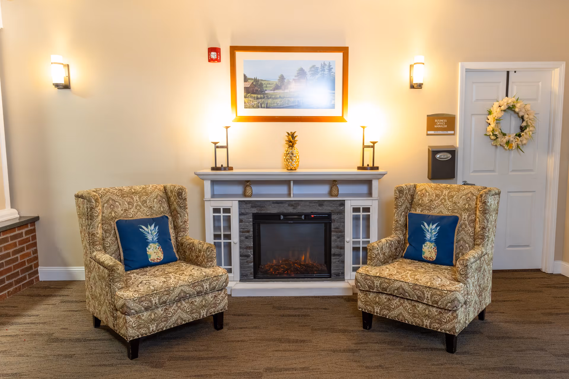 A cozy seating area with two patterned armchairs, each with a blue pillow featuring a pineapple design, positioned on either side of an electric fireplace. Above the fireplace is a framed landscape painting, and two lamps provide warm lighting. To the right, there is a white door decorated with a floral wreath and a sign that reads 'Business Office Manager'.