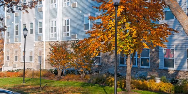 Exterior view of a multi-story senior living facility building with blue siding and stone accents. The scene includes autumn trees with orange and yellow leaves, green grass, and two black street lamps in the foreground.
