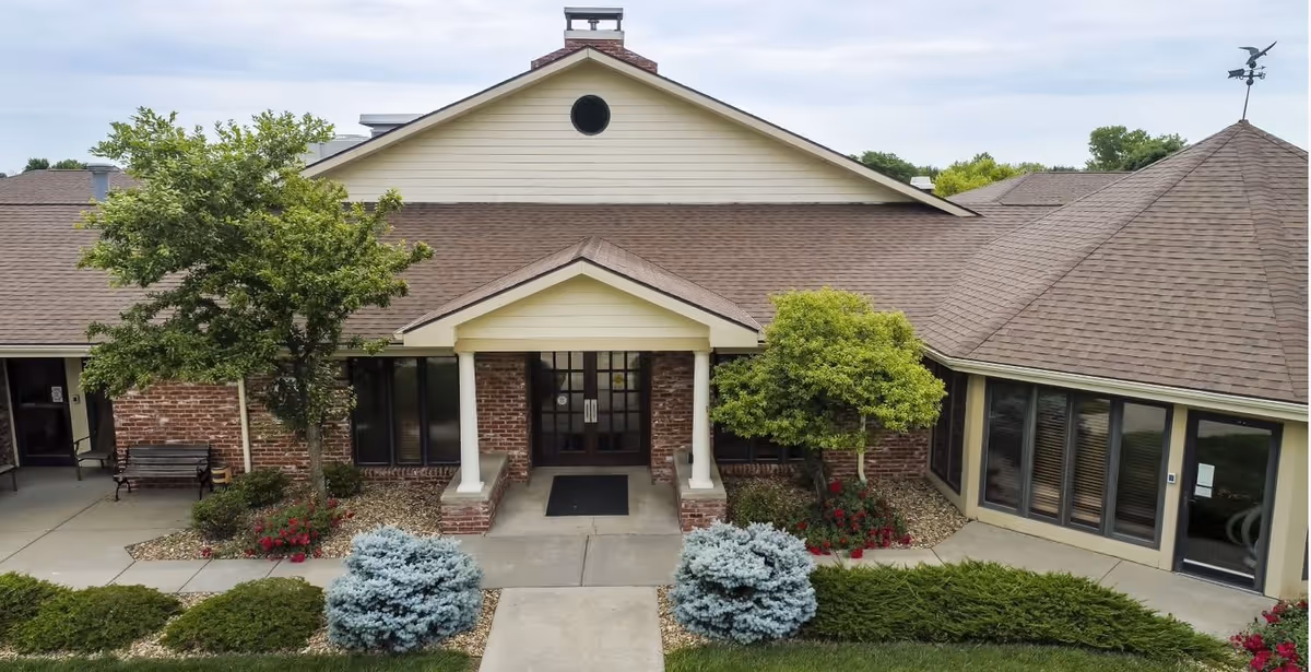 Front entrance of a single-story building with a brown shingled roof, brick and beige siding, two white columns supporting a small porch roof, surrounded by landscaped bushes, small trees, and flowers.