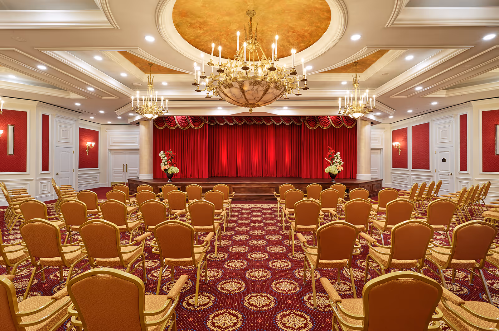 Ornate auditorium-style event room with rows of gold chairs facing a stage with red curtains and chandeliers.