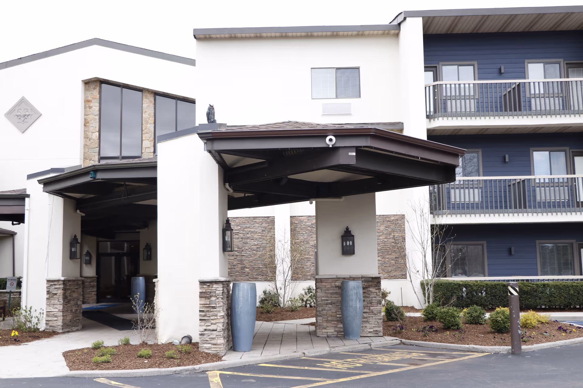 Entrance area of The Residences at Plainview by Chelsea Senior Living showing a covered drop-off with stone and white stucco pillars, large blue planters, and a building facade with balconies and windows.