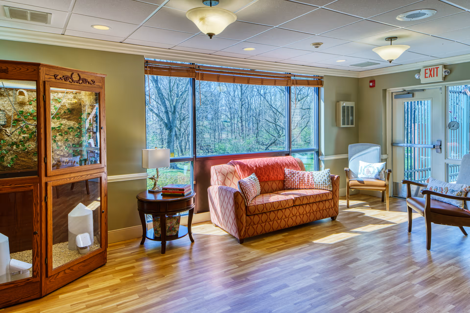 A bright and cozy living room area with a patterned orange loveseat adorned with three decorative pillows and a red throw blanket. Next to it is a round wooden side table with a lamp and a few books. Two armchairs with cushions are positioned near a glass door with an exit sign above it. Large windows behind the loveseat offer a view of leafless trees outside. The room has light wood flooring and soft green walls.