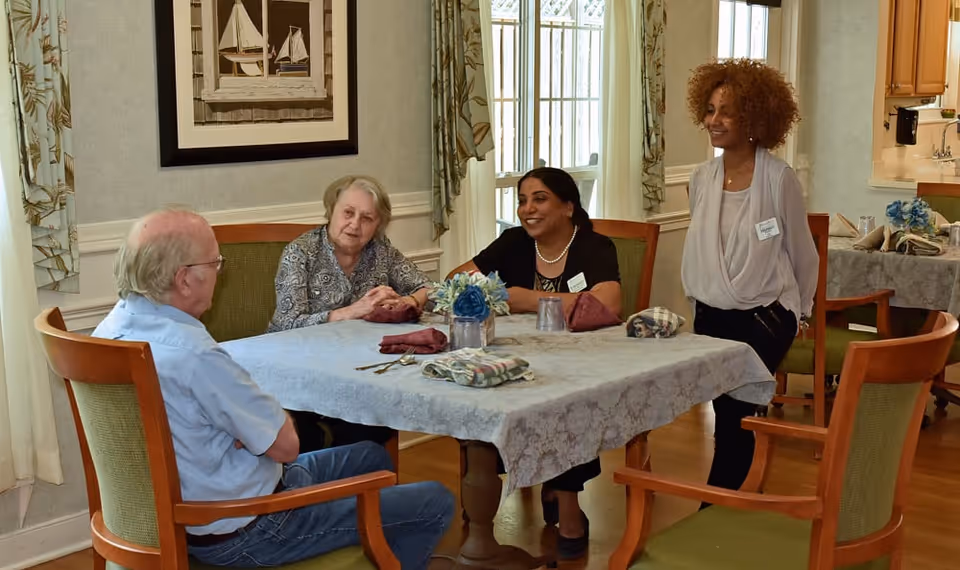 Three adults seated at a table and a standing staff member chatting in a senior living dining room.