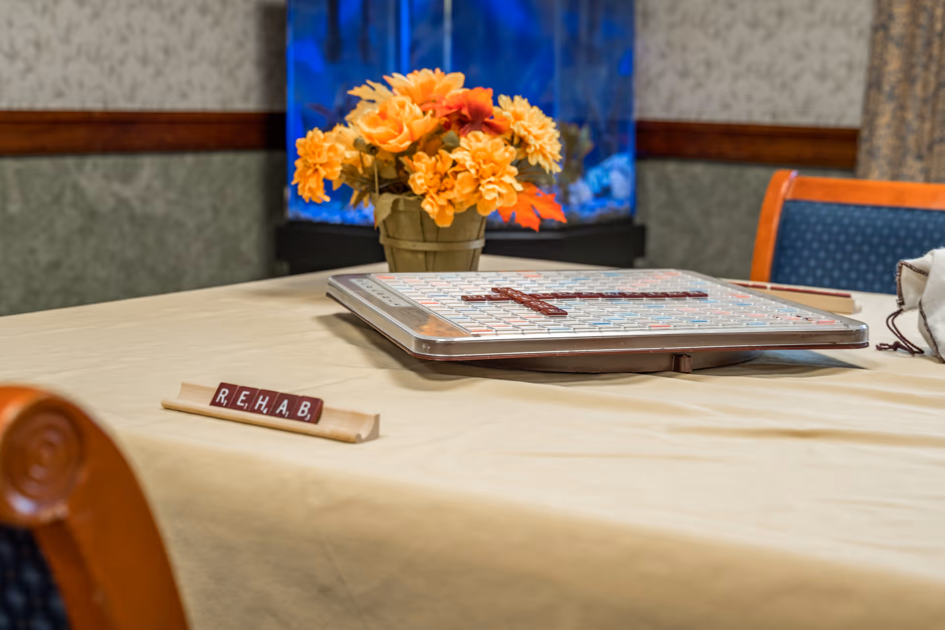 A table with a Scrabble game in progress, showing the word 'REHAB' on a tile rack. There is a vase with orange and yellow flowers on the table, and a blue aquarium in the background. The setting appears to be a cozy indoor common area with chairs around the table.