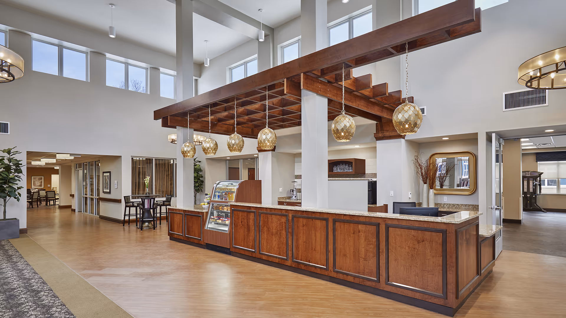 Spacious and well-lit interior area of a senior living facility with a large wooden reception or service counter featuring hanging decorative pendant lights. The room has high ceilings with multiple windows near the top, wood and granite finishes, and adjacent seating areas with tables and chairs. There are plants and a mirror on the walls, and the flooring is a combination of wood and carpet.