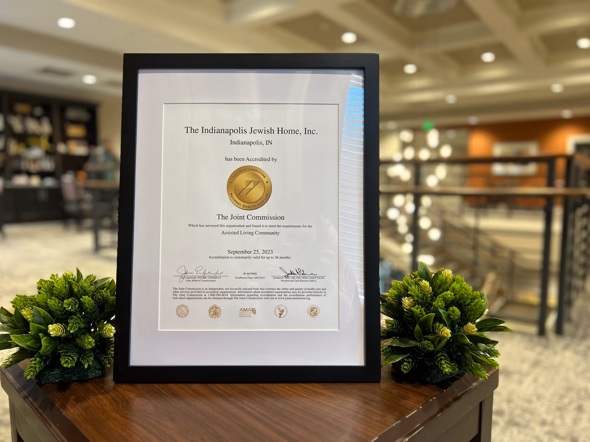 A framed accreditation certificate from The Joint Commission for The Indianapolis Jewish Home, Inc. displayed on a wooden table with two small green plants on either side. The background shows a blurred interior space with railings and lights.