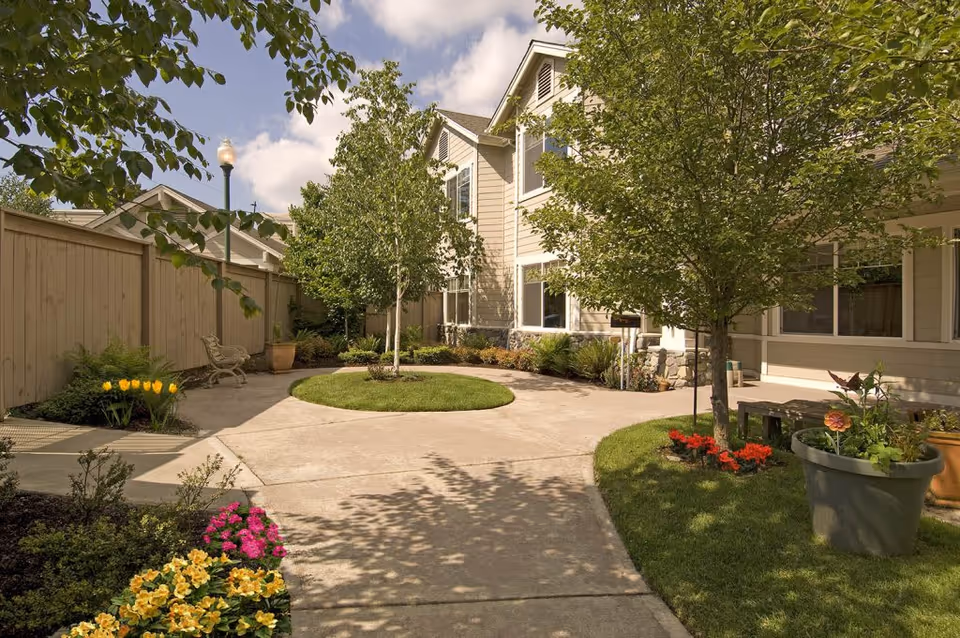 A sunny outdoor courtyard area at Aegis Living Callahan House featuring a paved walkway, green grass patches, several trees, colorful flower beds, a wooden fence, and a beige two-story building with multiple windows in the background.