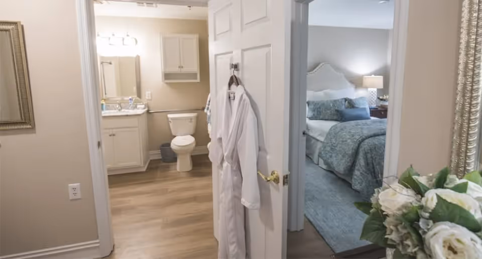Interior view of a hallway showing an open bathroom with a vanity and toilet on the left and a furnished bedroom with a bed and nightstand on the right, robes hanging on the door and a vase of flowers in the foreground.
