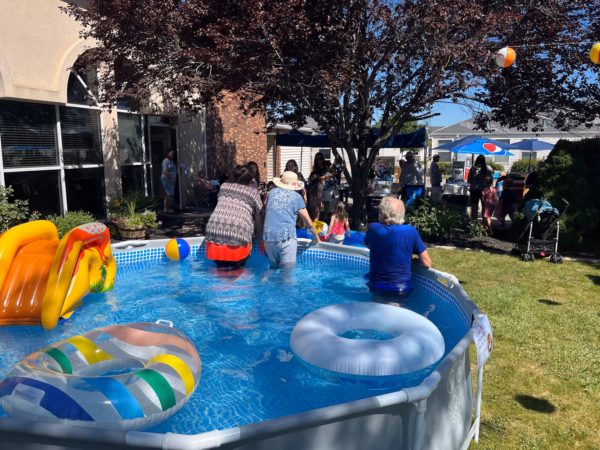 People enjoying a sunny day at an outdoor gathering at Country Pines Retirement Community. Several individuals are standing in a small above-ground pool with inflatable toys, while others are gathered under umbrellas and tents in the background near a building. There are trees providing shade and a stroller on the grass nearby.