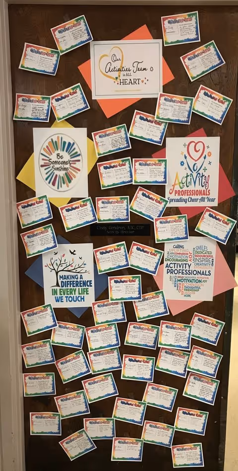 A bulletin board filled with multiple colorful cards and posters related to activities and activity professionals. The posters include motivational phrases such as 'Our Activities Team is All Heart,' 'Be Someone's Sunshine,' 'Making a Difference in Every Life We Touch,' and 'Activity Professionals Spreading Cheer All Year.' The cards appear to be activity schedules or notes, arranged neatly across the board.