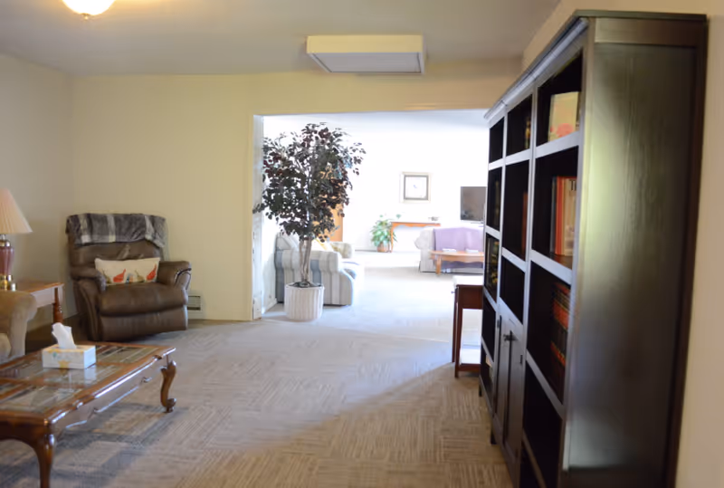Spacious living room in a senior facility with chairs, a coffee table, and a tall bookshelf.