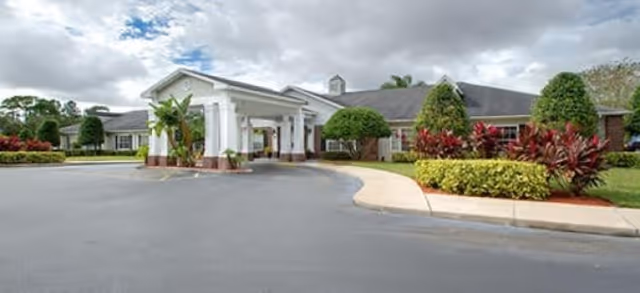 Exterior view of a single-story senior living facility with a covered entrance, surrounded by well-maintained landscaping including bushes and small trees under a cloudy sky.