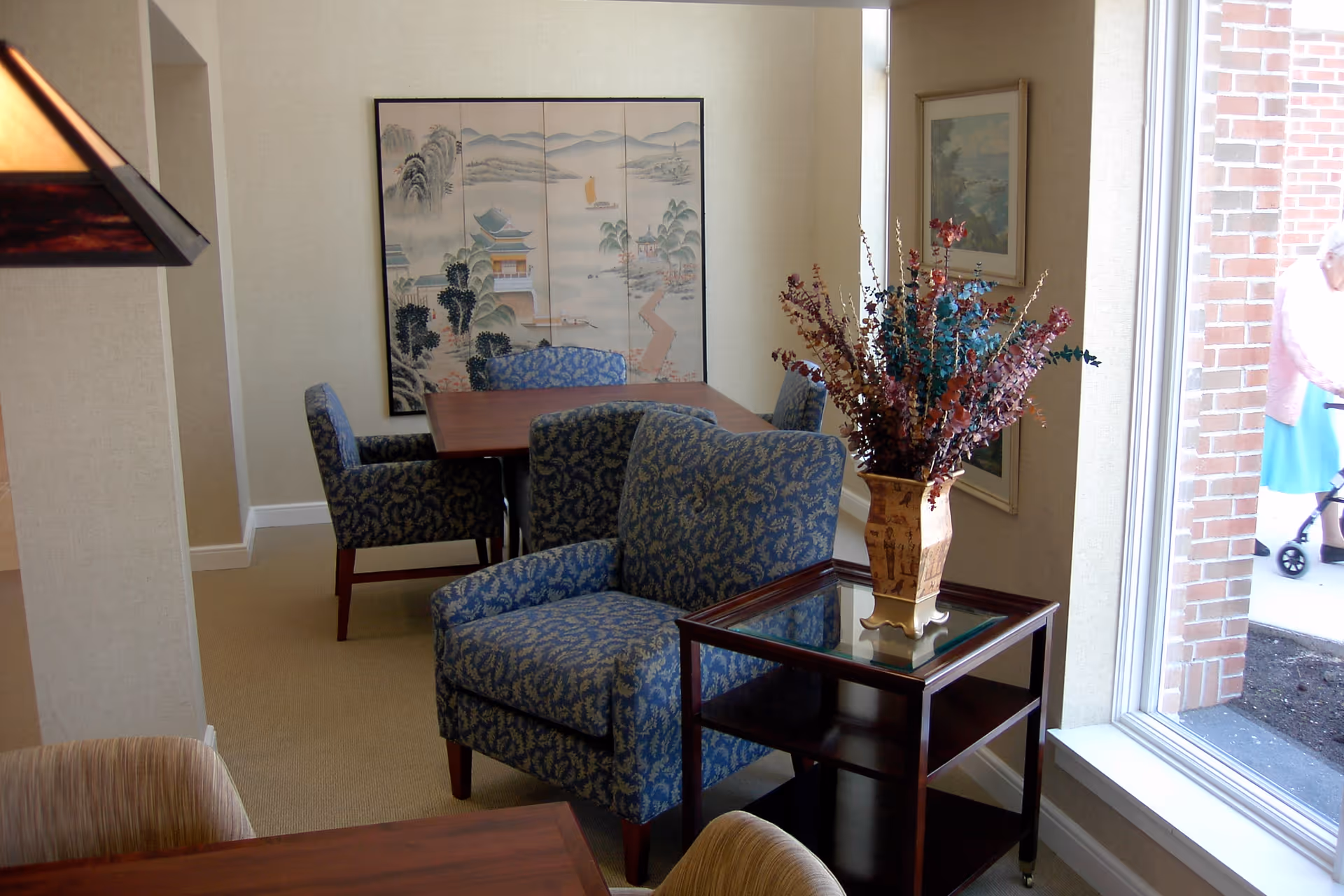 A cozy interior room with patterned blue armchairs and a wooden table. A decorative vase with dried flowers sits on a glass-topped side table near a large window. On the wall, there is a framed Asian-style painting and another framed artwork. Outside the window, a person with a walker is partially visible near a brick wall.