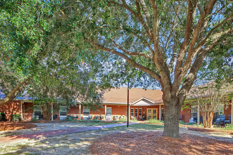 A single-story brick building with a brown shingled roof surrounded by large trees and a shaded courtyard area with a paved walkway and some outdoor seating.