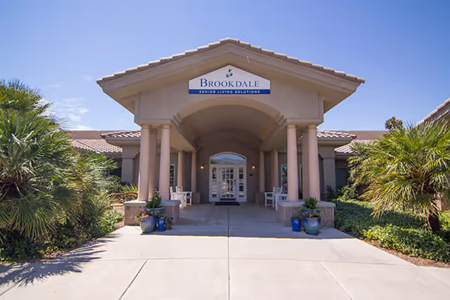 Front entrance of Brookdale Tanque Verde senior living facility with a covered driveway supported by columns, surrounded by greenery and palm trees under a clear blue sky.