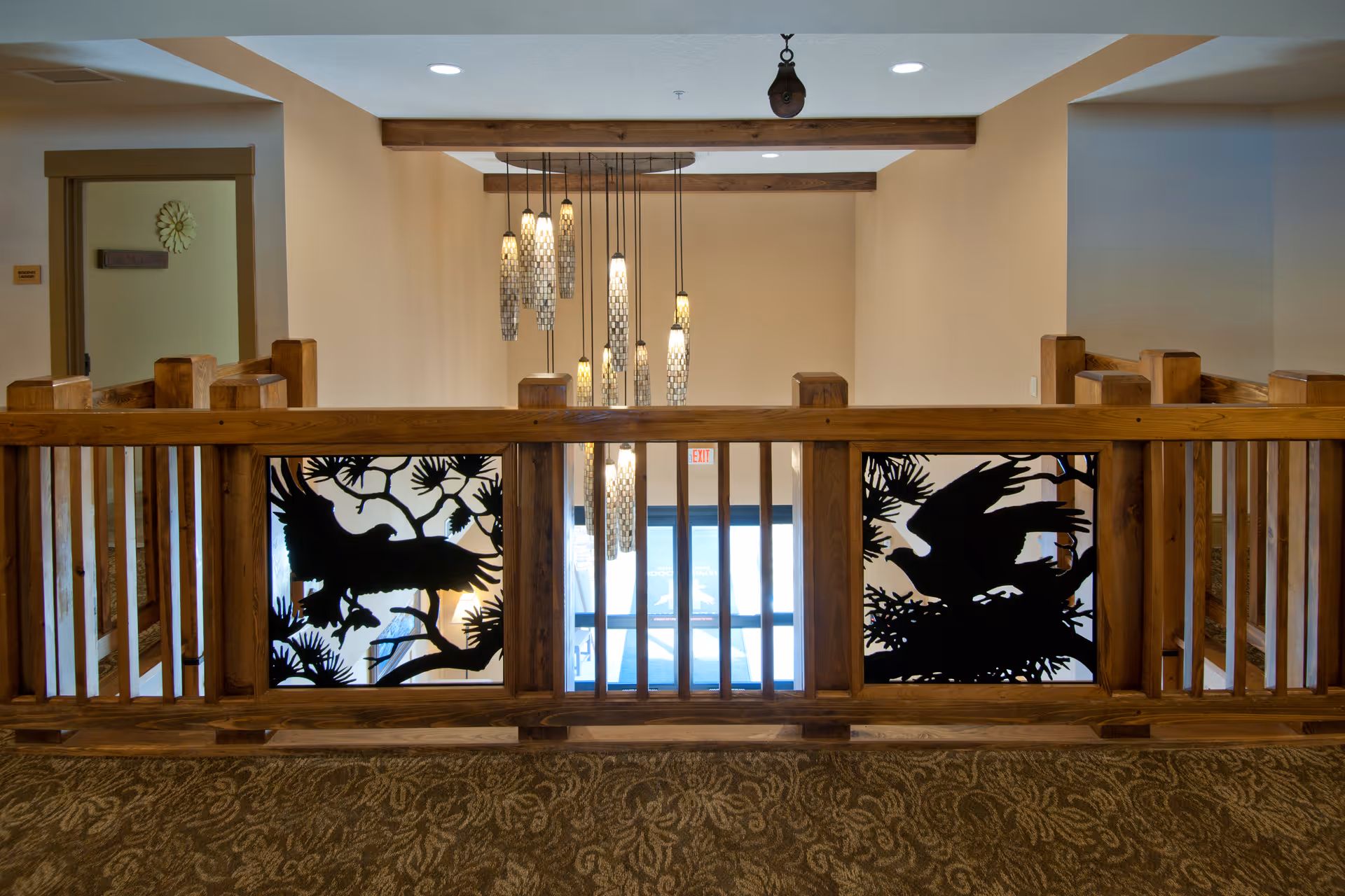 Interior view of a senior living facility hallway with wooden railing featuring decorative panels with bird silhouettes. A modern chandelier with multiple hanging lights is visible in the background, along with beige walls and a carpeted floor with a floral pattern.